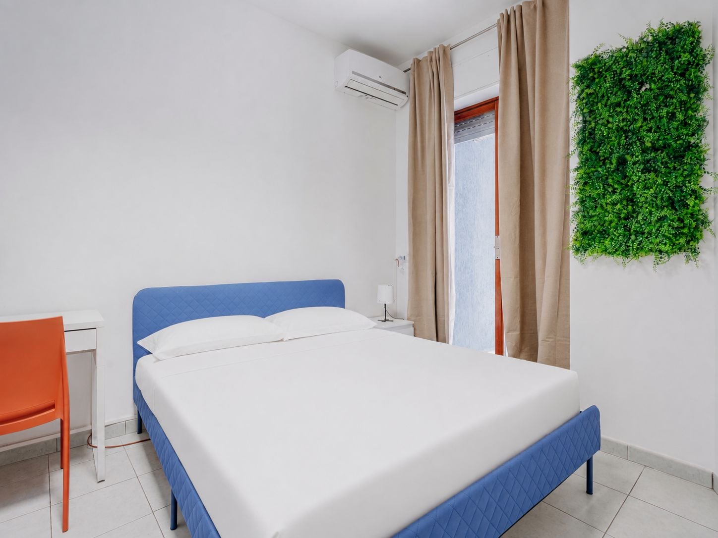 A bedroom with a bed, a desk, an air conditioner, and a green wall decoration.