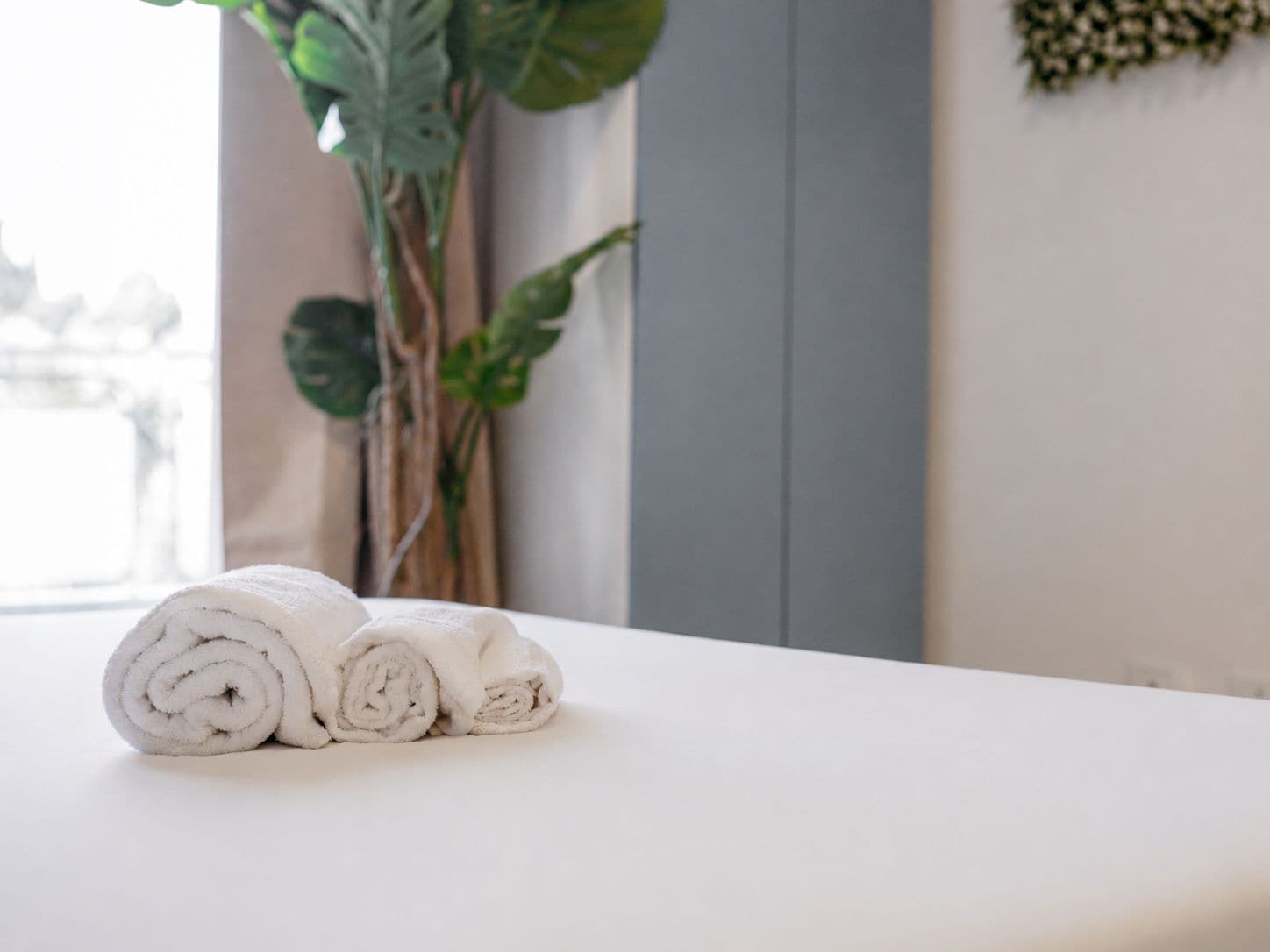 Een keurig opgerolde witte handdoek op een wit oppervlak, waarschijnlijk een badkamermeubel, met een grote groene plant op de achtergrond.