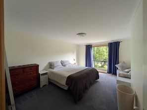 A bedroom with a bed, a dresser, a nightstand, a window with curtains, and a view of greenery outside.