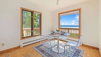 Un área de comedor con una mesa y sillas, ventanas amplias con vista a la playa y suelo de madera.