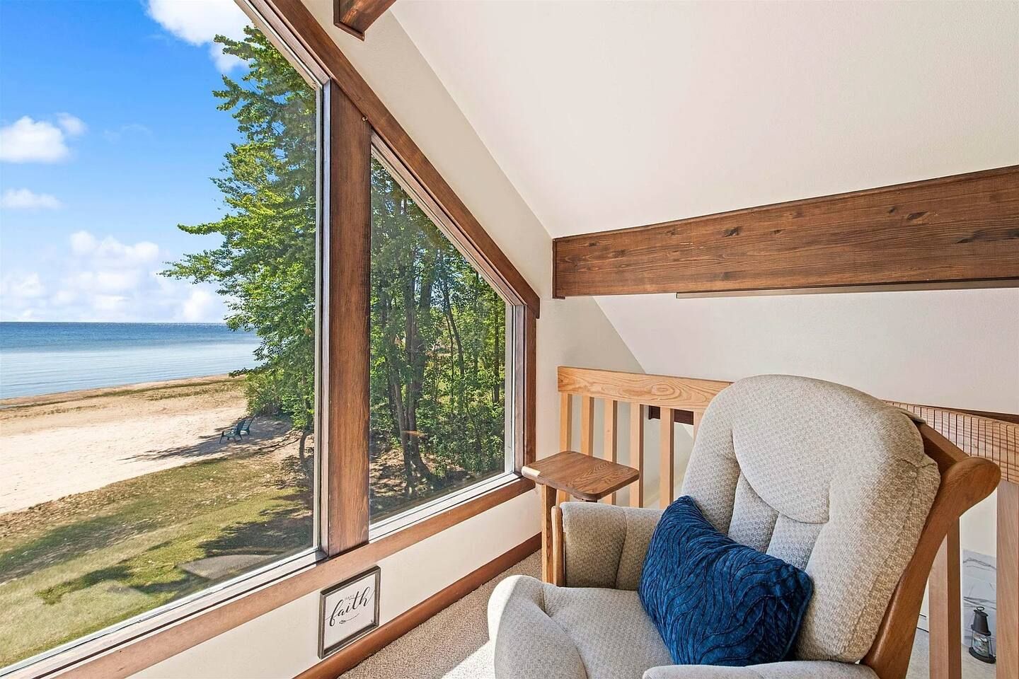 Une pièce dotée d’une grande fenêtre donnant sur une plage et une forêt, d’une chaise en bois avec des coussins et d’une petite table.