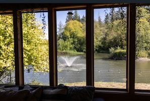 Quarto com grandes janelas dando para um lago com fonte, cercado por árvores e uma cerca.
