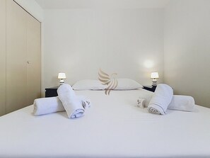 Ein Hotelzimmer mit einem ordentlich bezogenem Bett, zwei aufgerollten Handtüchern, einem Kopfkissen mit einer Feder-Applikation und zwei Nachttischlampen.