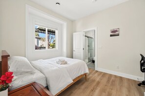 Un dormitorio con cama, ventana, puerta y un cuadro pequeño en la pared.