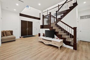 Amplia sala de estar con una escalera de madera, un sofá, un televisor sobre un mueble blanco y una puerta que conduce a otra habitación.