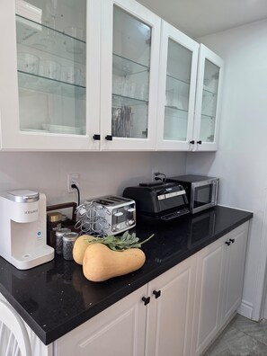 A kitchen with white cabinets, a black countertop, a coffee maker, a toaster, a microwave, and a glass cabinet with utensils. - Stay Cozy Orchard (Clarington)