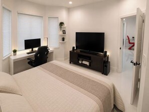 Une chambre à coucher avec un lit, un bureau avec un ordinateur, une télévision et une porte devant laquelle se trouve une chaise rouge.