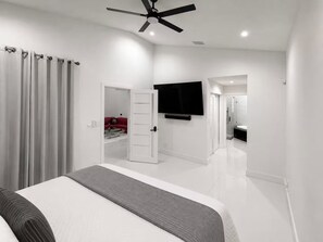 A modern bedroom with a bed, a ceiling fan, a TV, and a bathroom in the background. - Lakefront Pool Home near Wellington Polo & Beaches (West Palm Beach)