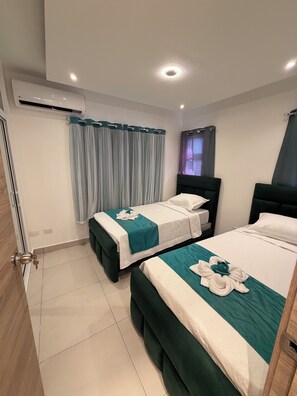 A hotel room with two beds, each with a teal bedspread and white pillows, a window with a curtain, and a door with a key in the lock. - MOONLIGHT APARTMENT NEAR THE BEACH (Puerto Plata)