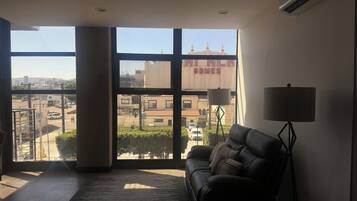 A living room with a large window, a black leather sofa, a floor lamp, and a view of a building with a sign reading 'HI HLA GAMES'.