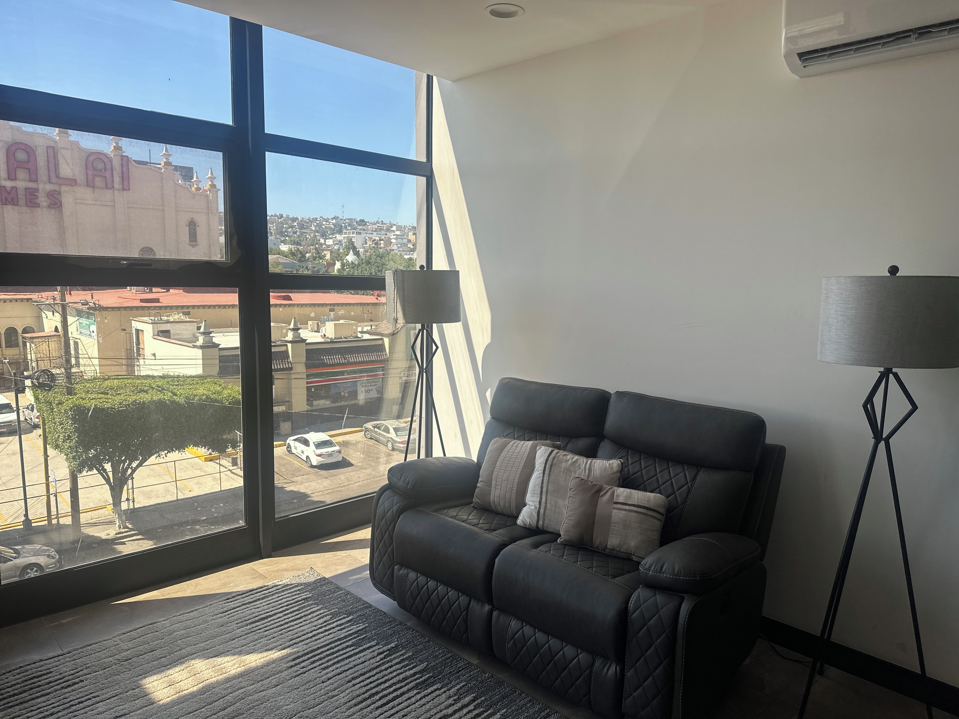 A room with a large window, a black leather sofa, a floor lamp, and a view of a building with 'ALAI' signage.