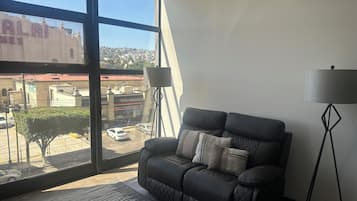 A room with a large window, a black leather sofa, a floor lamp, and a view of a building with 'ALAI' signage.