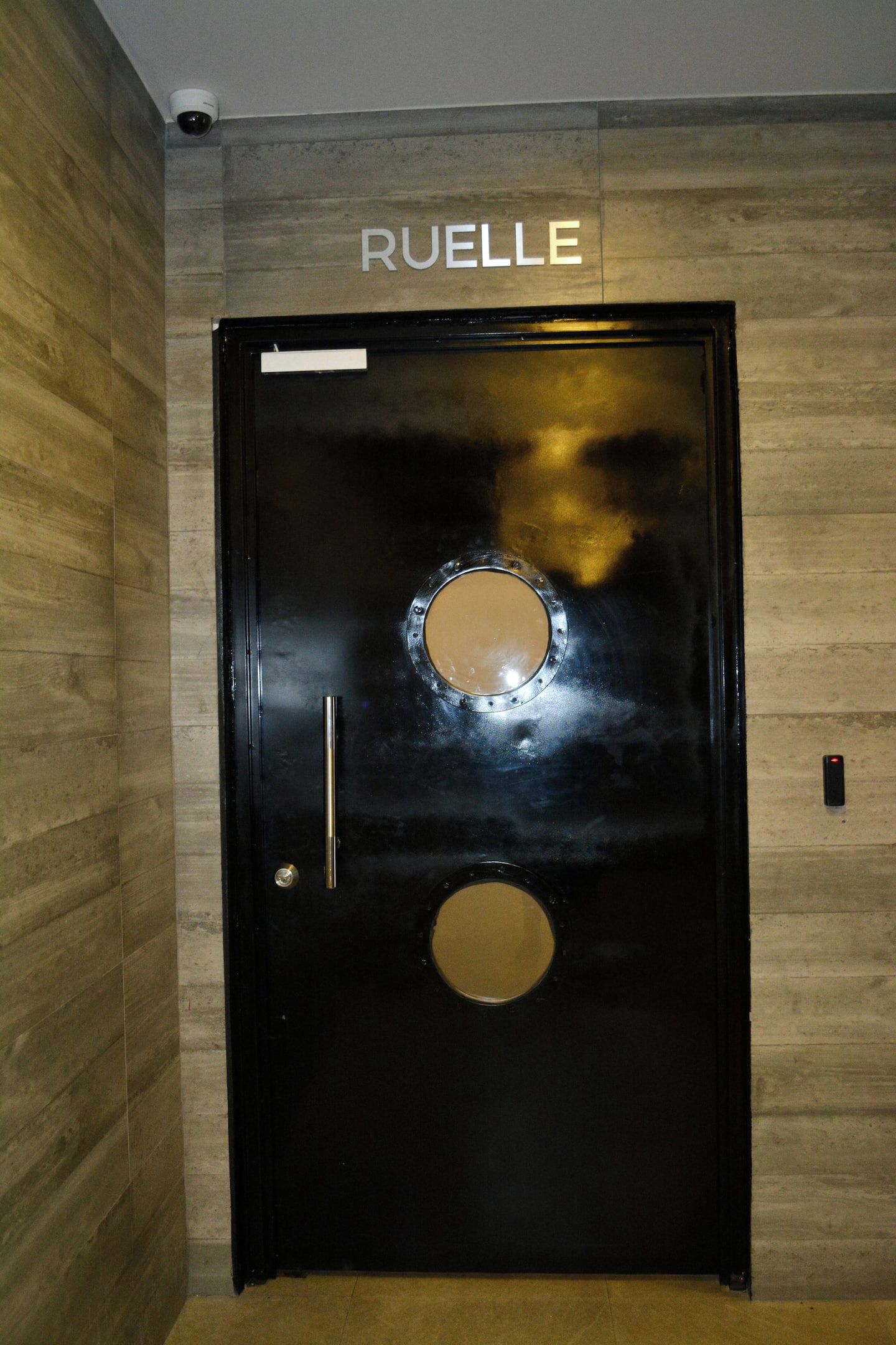 Una puerta negra con la palabra 'RUELLE' encima, una ventana redonda y una cerradura.