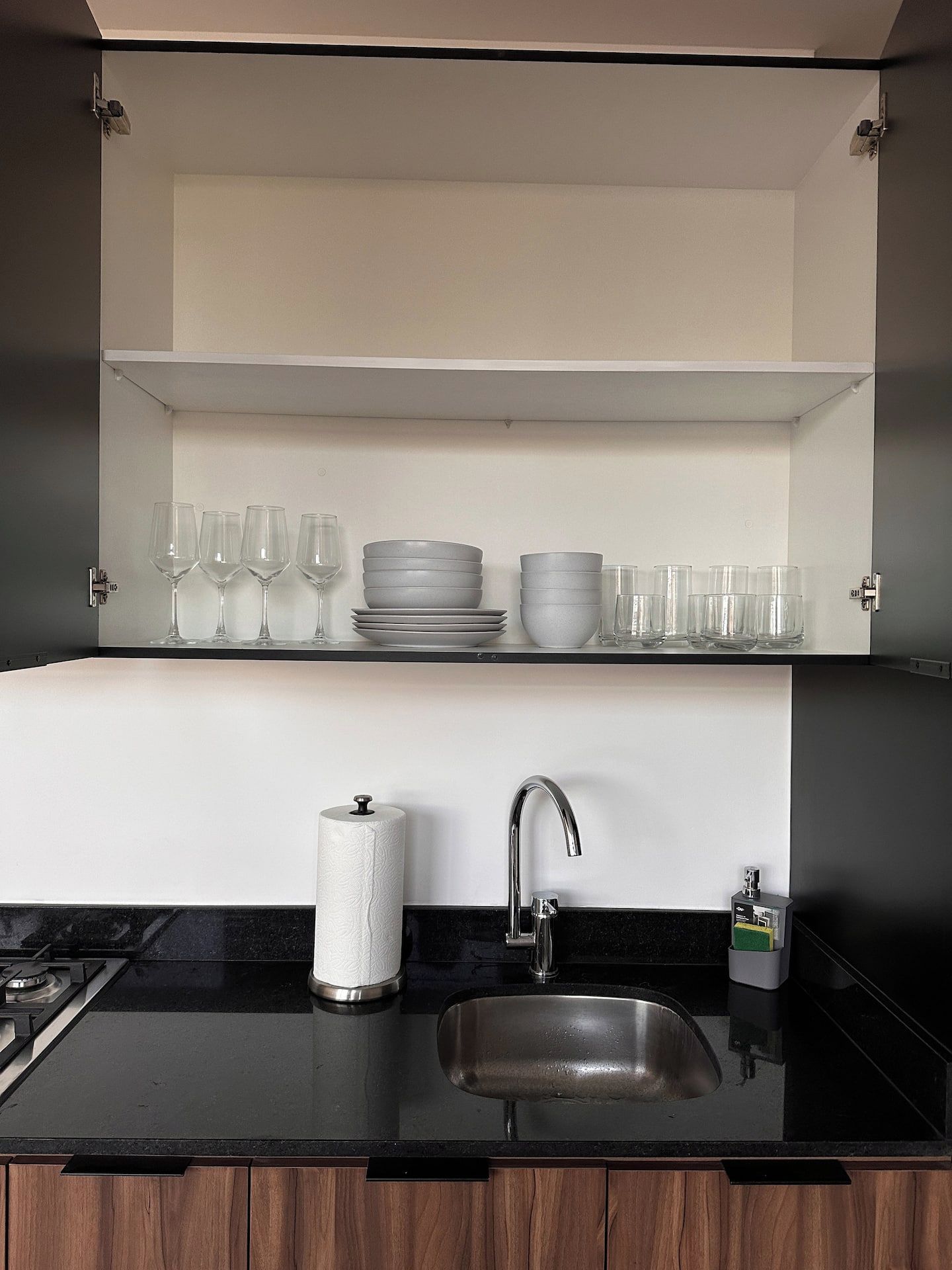 A kitchen with a black countertop, a stainless steel sink, a paper towel holder, and a shelf with stacked plates and glasses.