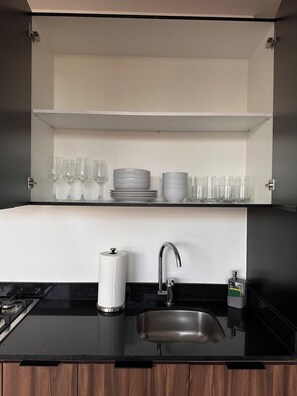 A kitchen with a black countertop, a stainless steel sink, a paper towel holder, and a shelf with stacked plates and glasses.