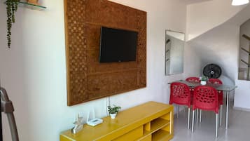 A modern living room with a flat-screen TV mounted on a textured wall, a yellow entertainment unit, red chairs around a glass table, and a ceiling fan.