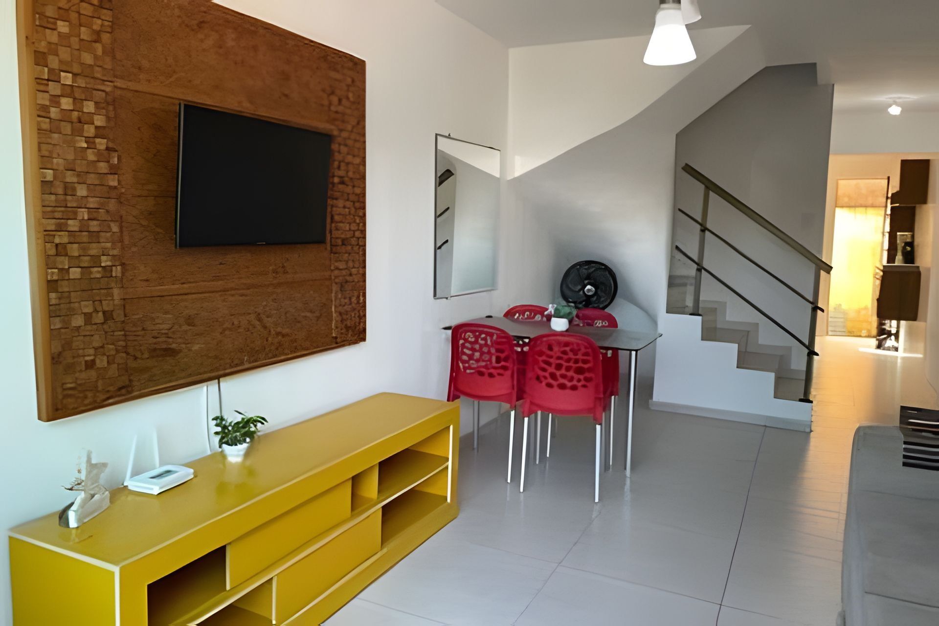Un salon moderne avec un téléviseur à écran plat fixé à un mur en briques, un meuble de rangement jaune, un coin repas avec des chaises rouges et un escalier menant à un autre niveau.