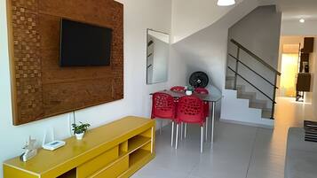 Un salon moderne avec un téléviseur à écran plat fixé à un mur en briques, un meuble de rangement jaune, un coin repas avec des chaises rouges et un escalier menant à un autre niveau.