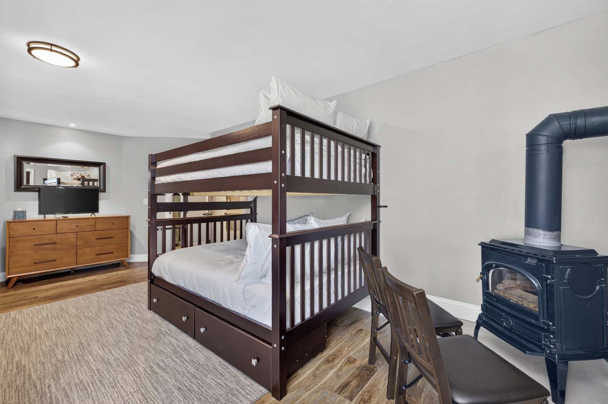 A bunk bed with a wooden frame and white bedding, a wooden dresser with drawers, a television on a stand, and a black wood-burning stove.