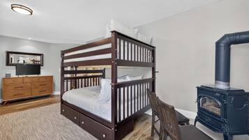 A bunk bed with a wooden frame and white bedding, a wooden dresser with drawers, a television on a stand, and a black wood-burning stove.