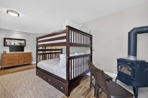 Una cama litera con estructura de madera y ropa de cama blanca, un aparador de madera con cajones, un televisor sobre un mueble bajo y un estufa de leña negra.