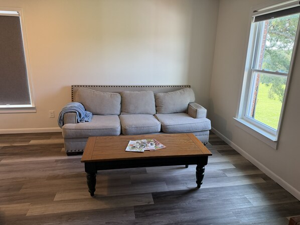 Una sala de estar con un sofá gris, una mesa de centro de madera y un ventanal con vista a follaje.