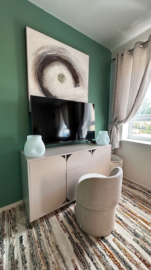 A room with a television on a white cabinet, a large abstract painting above it, a window with curtains, and a patterned rug.