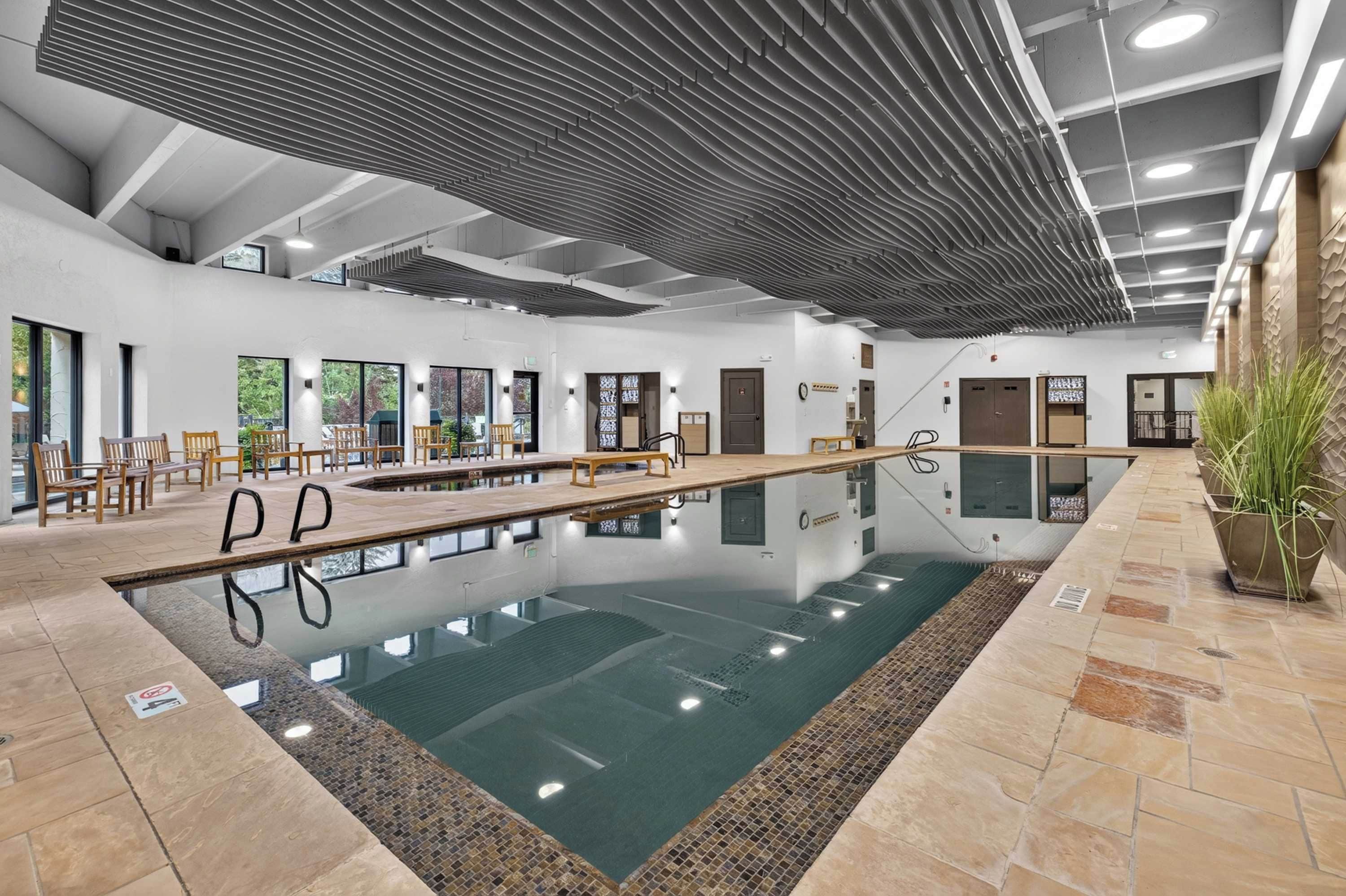 Ein Schwimmbecken mit Holzterrasse und Liegestühlen, umgeben von Steinfliesen und großen Fenstern.