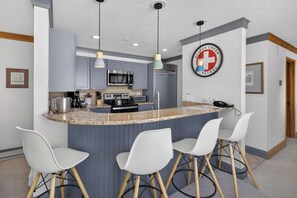 A modern kitchen with a granite countertop, stainless steel appliances, and bar stools. - Charter Ski In Ski Out 2BR (Avon)