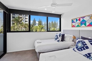 A bedroom with two beds, a ceiling fan, and a large window with a view of trees and buildings. - 2 Bedroom Luxury in Burleigh Heads! (Gold Coast)