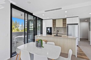 A modern kitchen with a central island, a dining area with a white table and chairs, and a view of the outdoors through a glass door. - 2 Bedroom Luxury in Burleigh Heads! (Gold Coast)