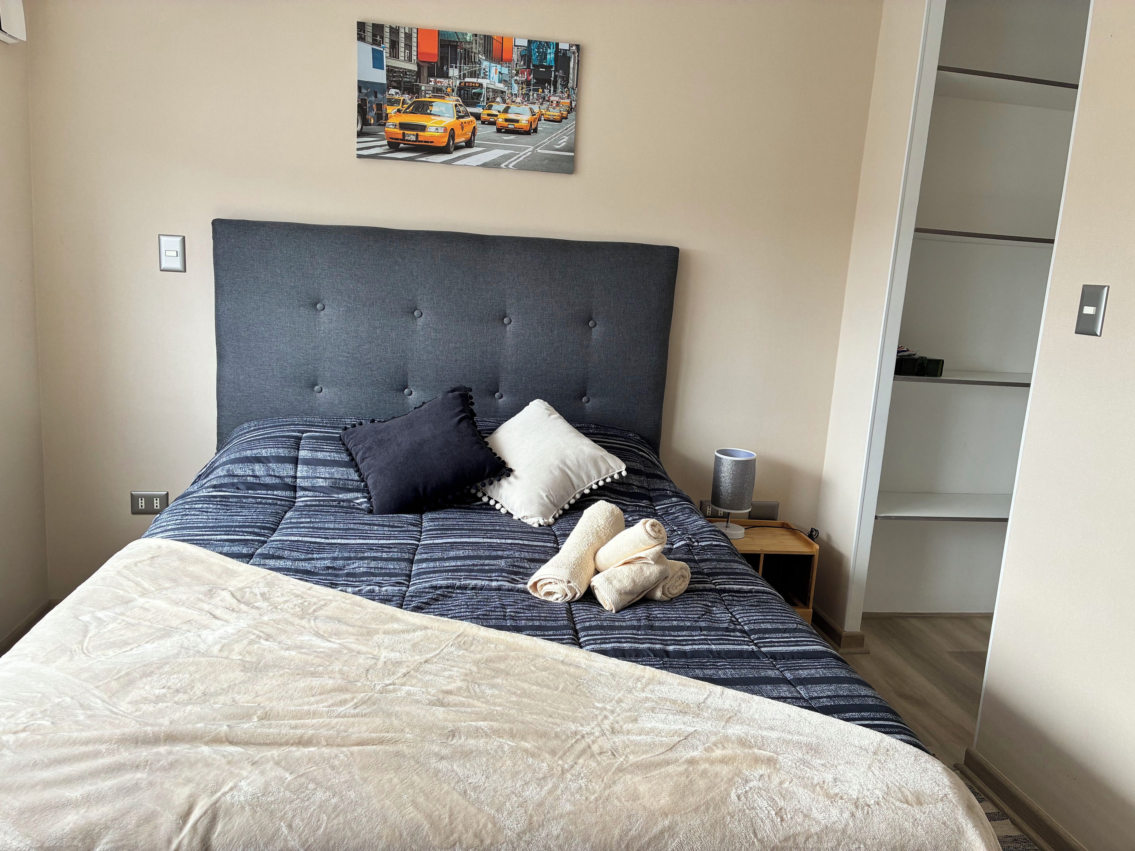 Una cama bien hecha con cabecera gris, dos almohadas y una toalla enrollada.