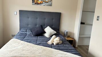 Una cama bien hecha con cabecera gris, dos almohadas y una toalla enrollada.