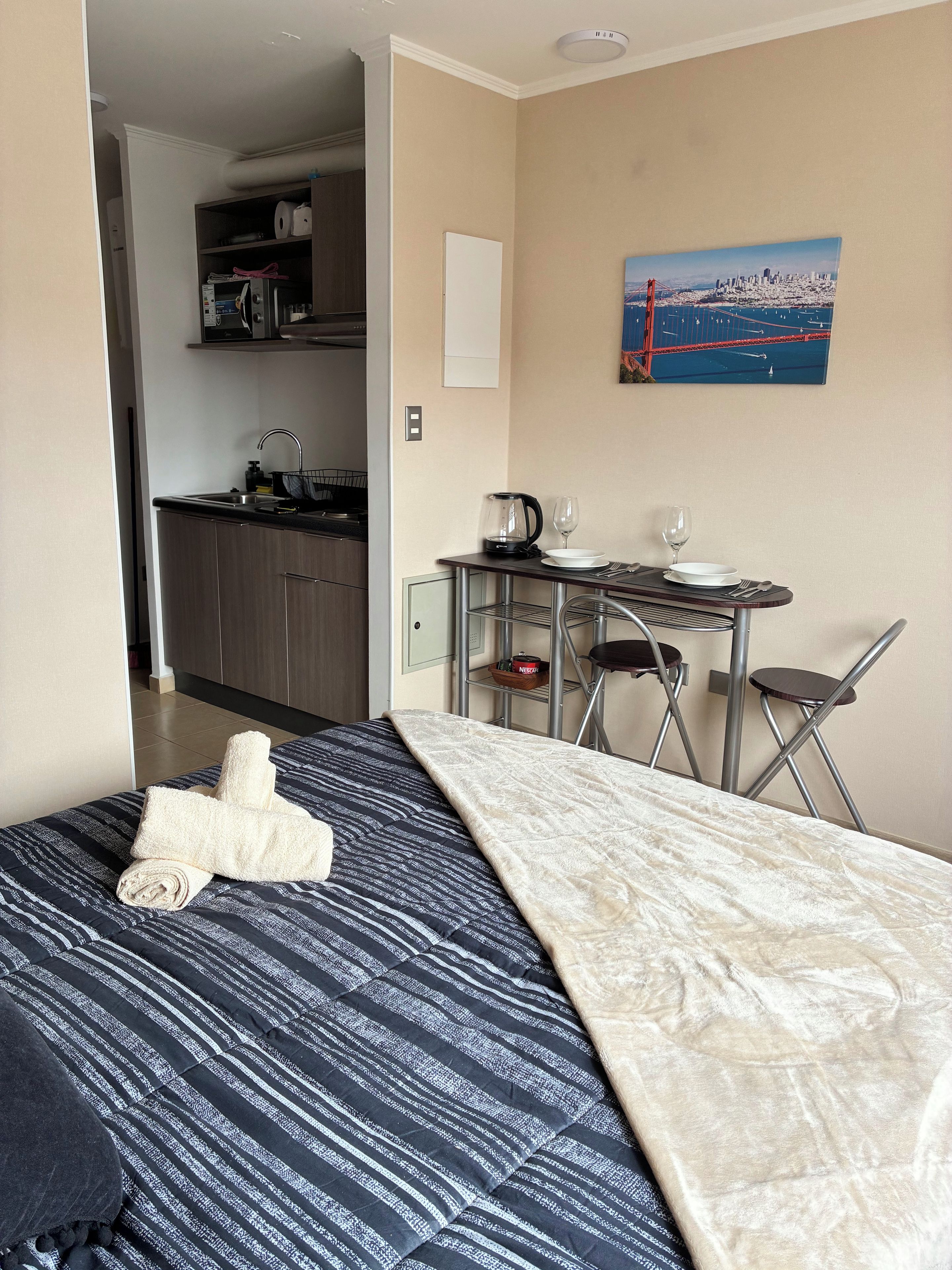 A hotel room with a bed, a small kitchenette, a dining table with chairs, and a painting on the wall.