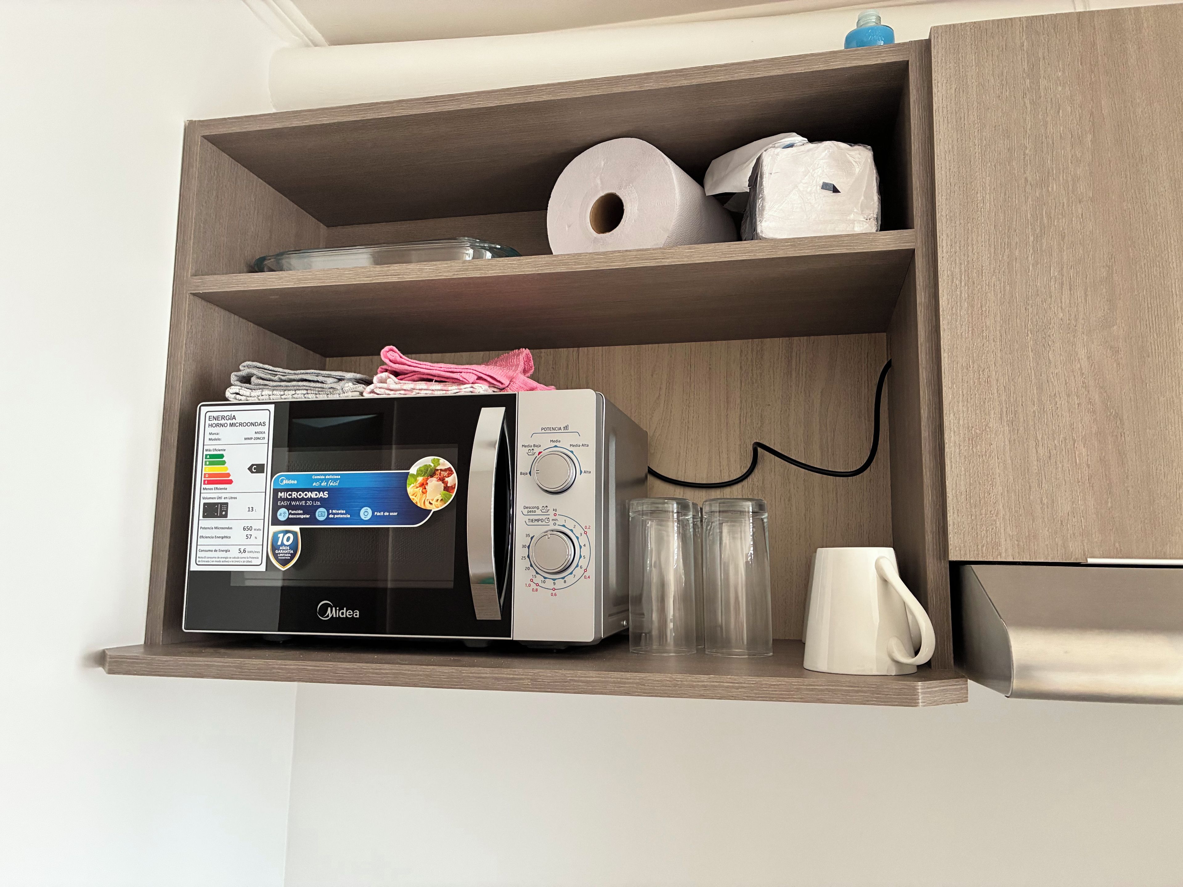 Una cocina con un microondas en un estante de madera, un rollo de toallas de papel y una taza blanca.