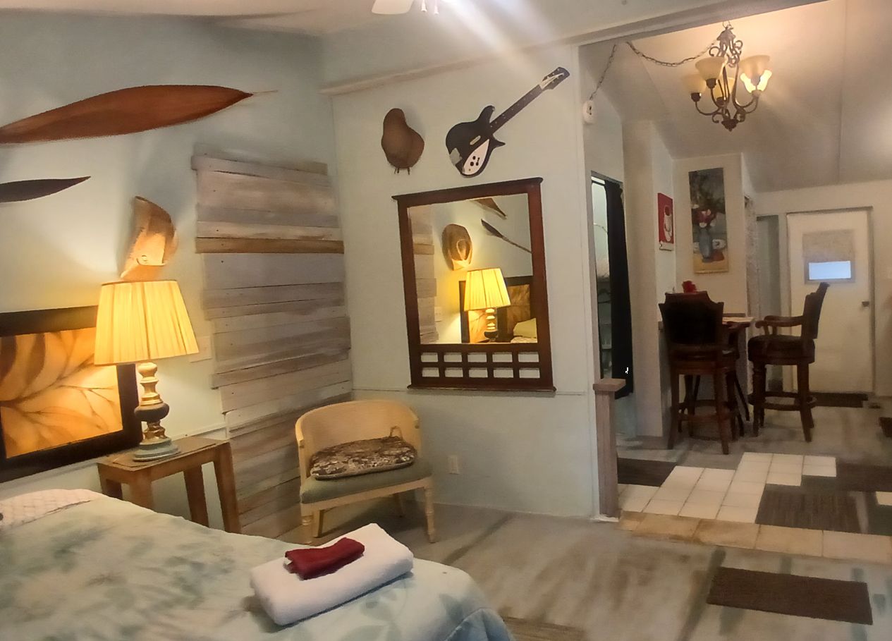 Ein Hotelzimmer mit Bett, Sessel, Esstisch mit Stühlen, Spiegel und Gitarre an der Wand.