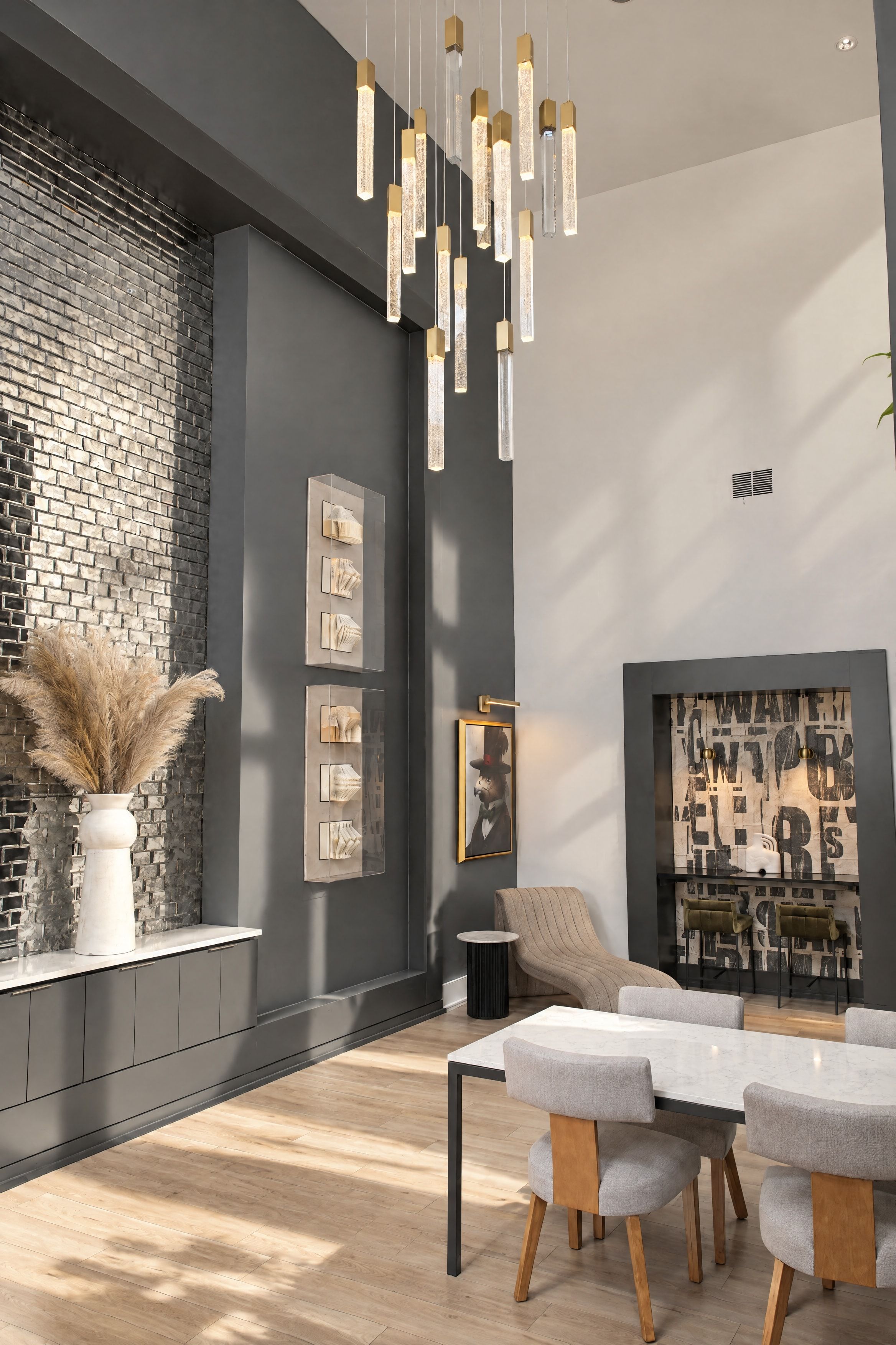 A modern interior with a brick wall, hanging pendant lights, a vase with pampas grass, framed artwork, and a seating area with a table and chairs.