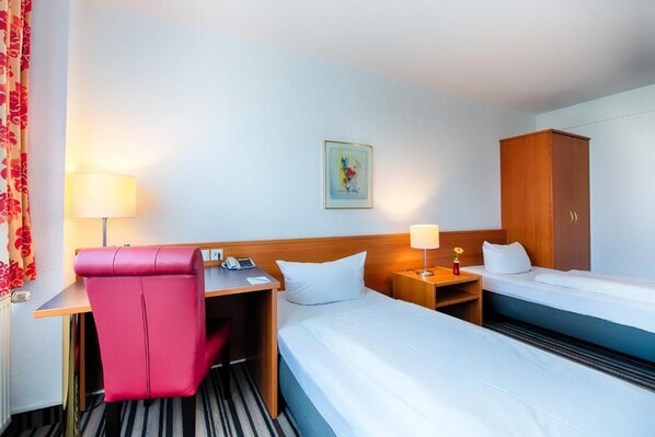 Ein Hotelzimmer mit Schreibtisch, Stuhl, Bett, Kleiderschrank und Nachttisch mit Lampe.