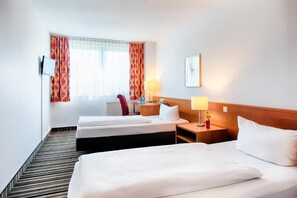 Ein Hotelzimmer mit zwei Betten, einem Schreibtisch mit Lampe, einem Stuhl und einem Fenster mit Vorhängen.