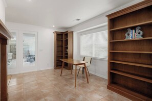 Une pièce comprenant une table à manger, des chaises, une bibliothèque intégrée et une fenêtre avec des stores.