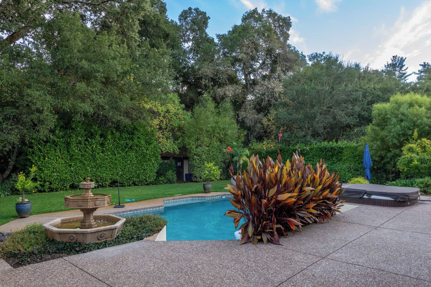 A swimming pool surrounded by lush greenery, a stone fountain, and a large potted plant.