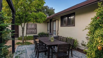 Un patio exterior con mesa y sillas, rodeado de follaje y un edificio con tejado marrón.