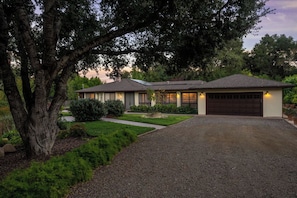A two-story house with a driveway leading to a garage, surrounded by mature trees and well-maintained landscaping.