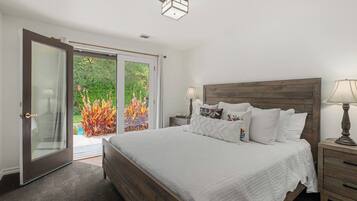 Une chambre dotée d’un grand lit, d’une tête de lit en bois, d’une table de chevet avec une lampe et d’une porte-fenêtre coulissante donnant sur un jardin.