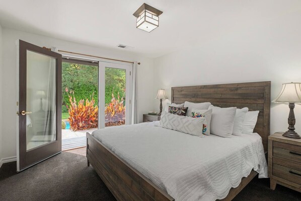 A bedroom with a large bed, a wooden headboard, a nightstand with a lamp, and a sliding glass door leading to a garden.