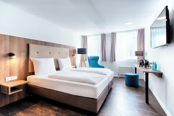 Ein modernes Hotelzimmer mit einem großen Bett, einem Schreibtisch mit Flachbildfernseher, einem Sessel und einem kleinen Hocker.