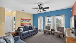 Ein Wohnzimmer mit einem blauen Sofa, gemusterten Sesseln, einem Deckenventilator und einem Fernseher.