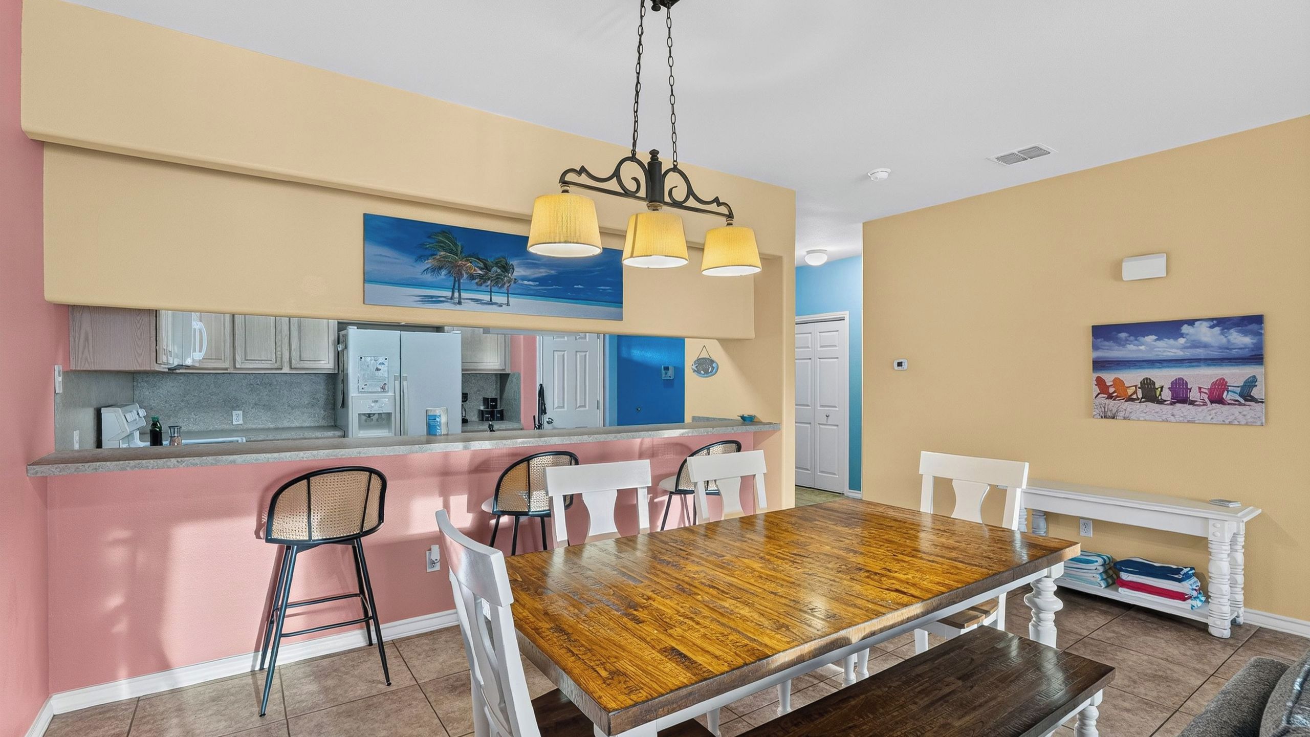 A dining area with a wooden table and chairs, a kitchen with white cabinets, and a beach-themed painting on the wall.