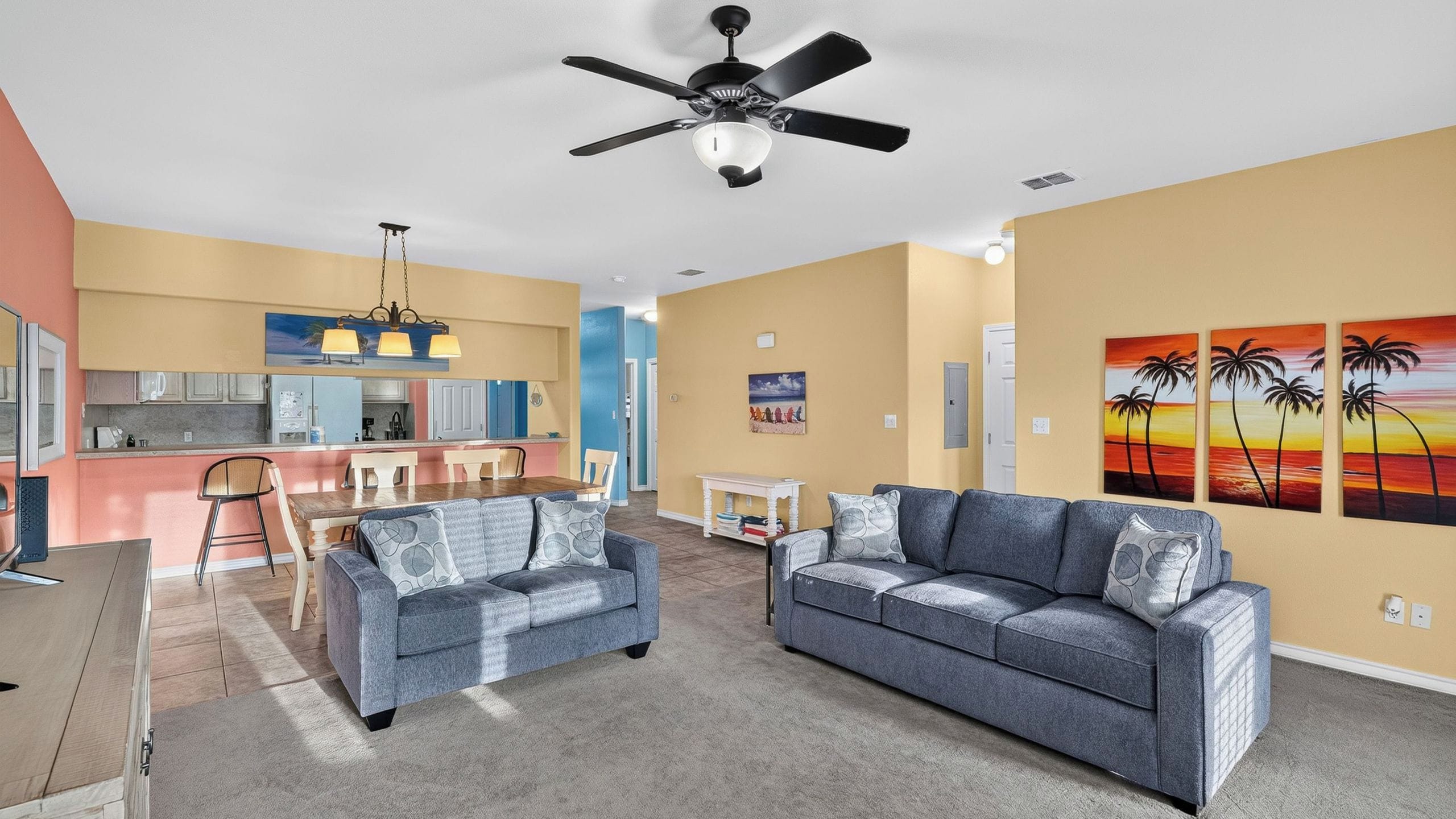 A living room with a grey sofa, a dining area with a pink table, and a kitchen with blue cabinets.