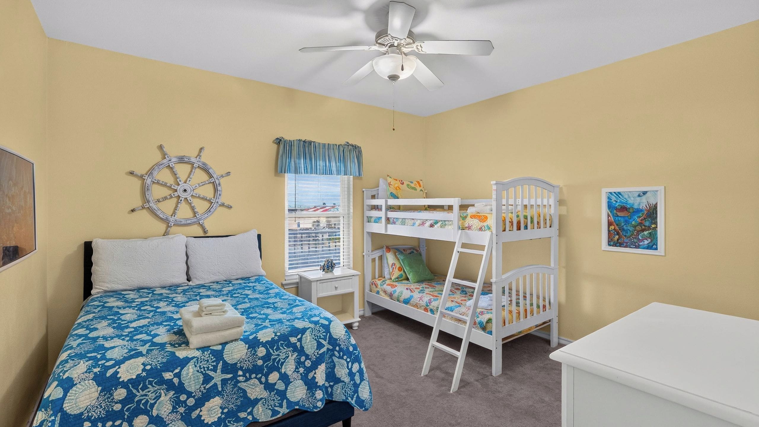 Een kinderkamer met stapelbedden, een bed met een blauw bloemenbedekking, een plafondventilator en een raam met jaloezieën.